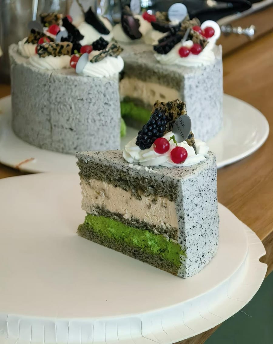 Una fetta di torta a strati: sesamo nero fuori, crema al centro, pan di Spagna verde alla base, con mora e ribes sulla panna.