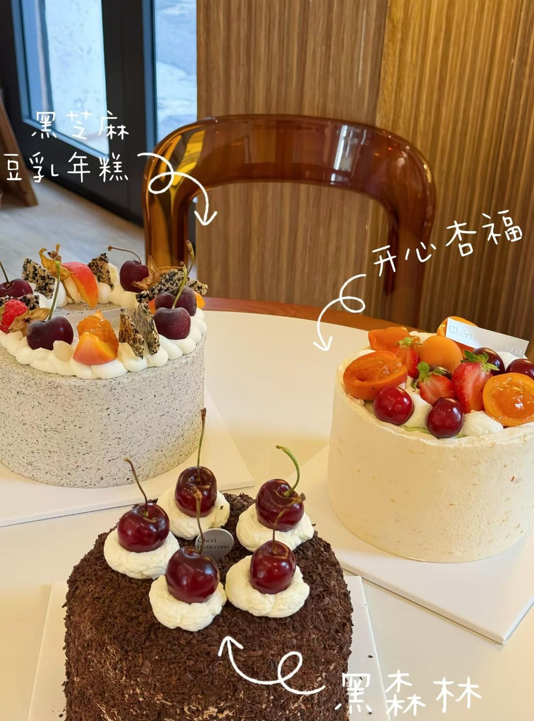 Tre torte intere su un tavolo bianco: una chiara punteggiata di sesamo nero con ciliegie, pesca e fiori secchi, una alla panna con albicocche, fragole e ciliegie, e una foresta nera coperta di scaglie di cioccolato. Sulla foto ci sono i loro nomi, scritti a mano in cinese.